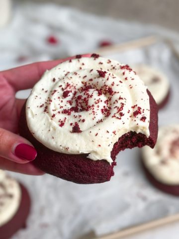 hand holding red velvet cookie with cream cheese frosting on top with a bite taken out of it
