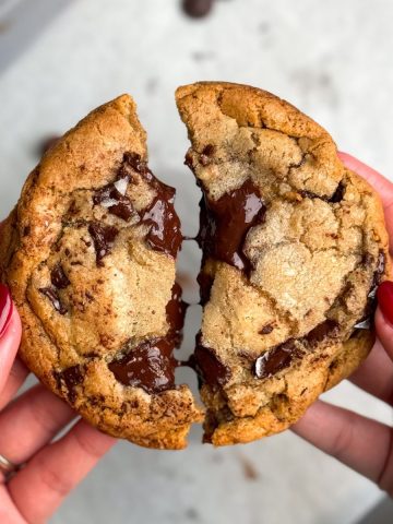 hand holding two halves of a large chocolate chip cookie with puddles of melted chocolate and flaky sea salt sprinkled on top