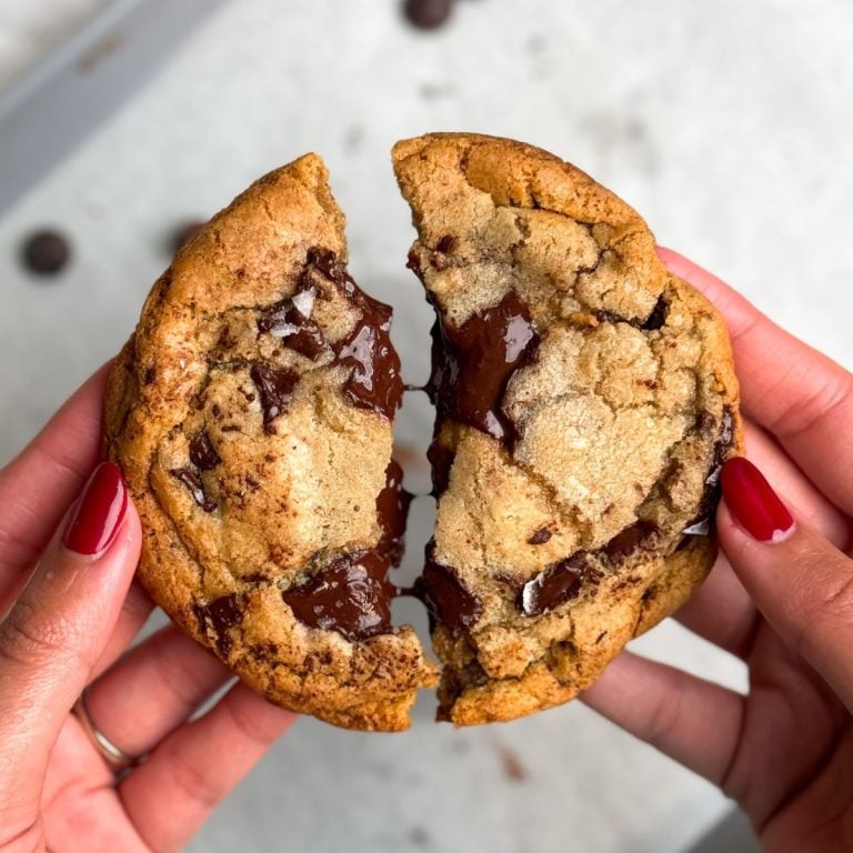 hand holding two halves of a large chocolate chip cookie with puddles of melted chocolate and flaky sea salt sprinkled on top