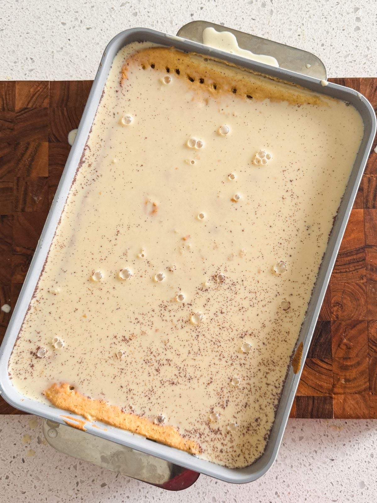 A rectangular baking dish filled with a pale yellow, creamy liquid over a tres leches cake base. The surface is sprinkled with cinnamon and has small bubbles, with some mixture spilled on the edges and countertop below.