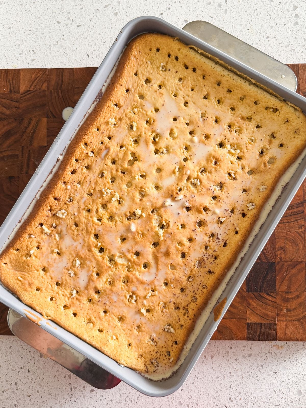 A rectangular yellow tres leches cake in a baking pan with holes poked across the top and glaze drizzled over it, set on a wooden cutting board and white countertop.