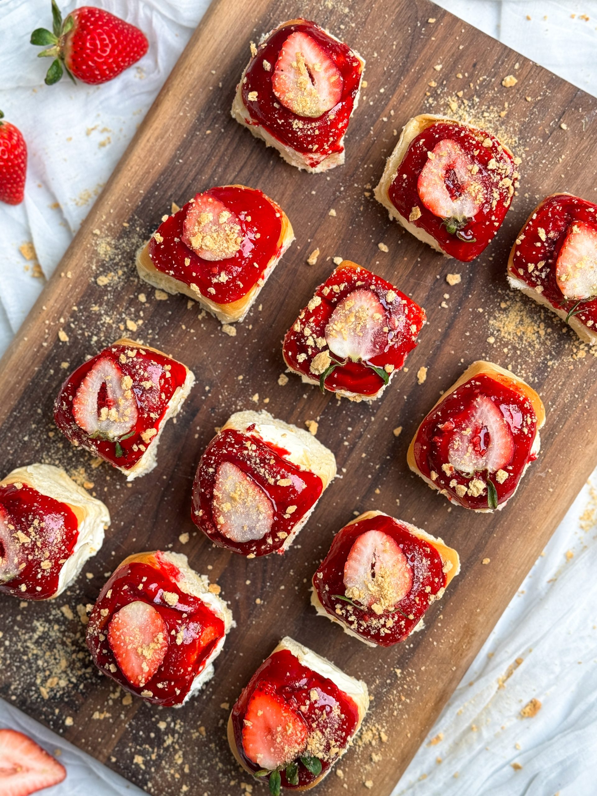 12 strawberry cheesecake buns on a wooden cutting board. buns are filled with a cheesecake filling, topped with a shiny strawberry glaze, and decorated with strawberries and crushed graham crackers. picture from the top