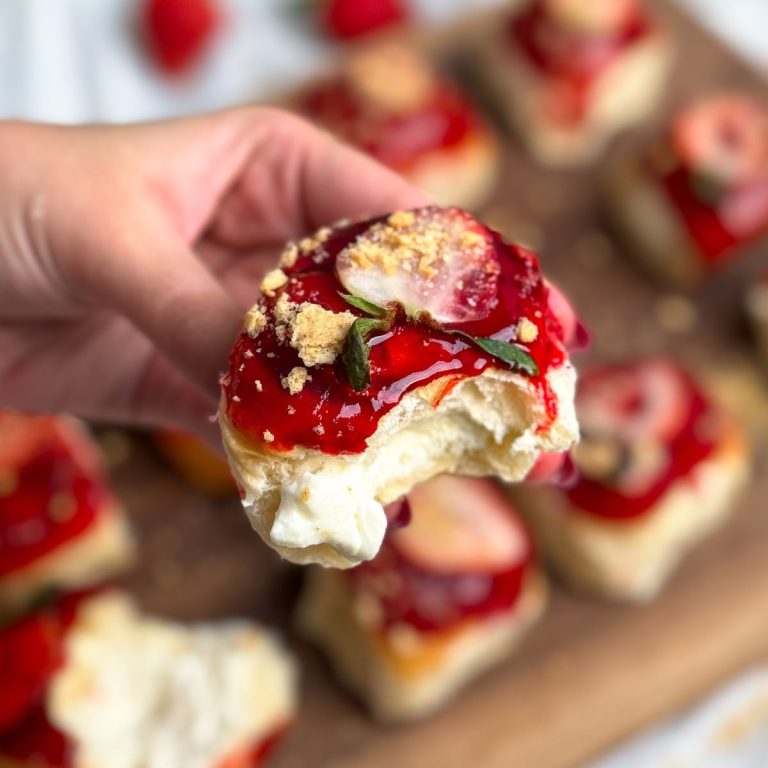 hand holding a strawberry cheesecake bun with a bite taken out to show the cheesecake filling inside. bun has a shiny strawberry glaze on top