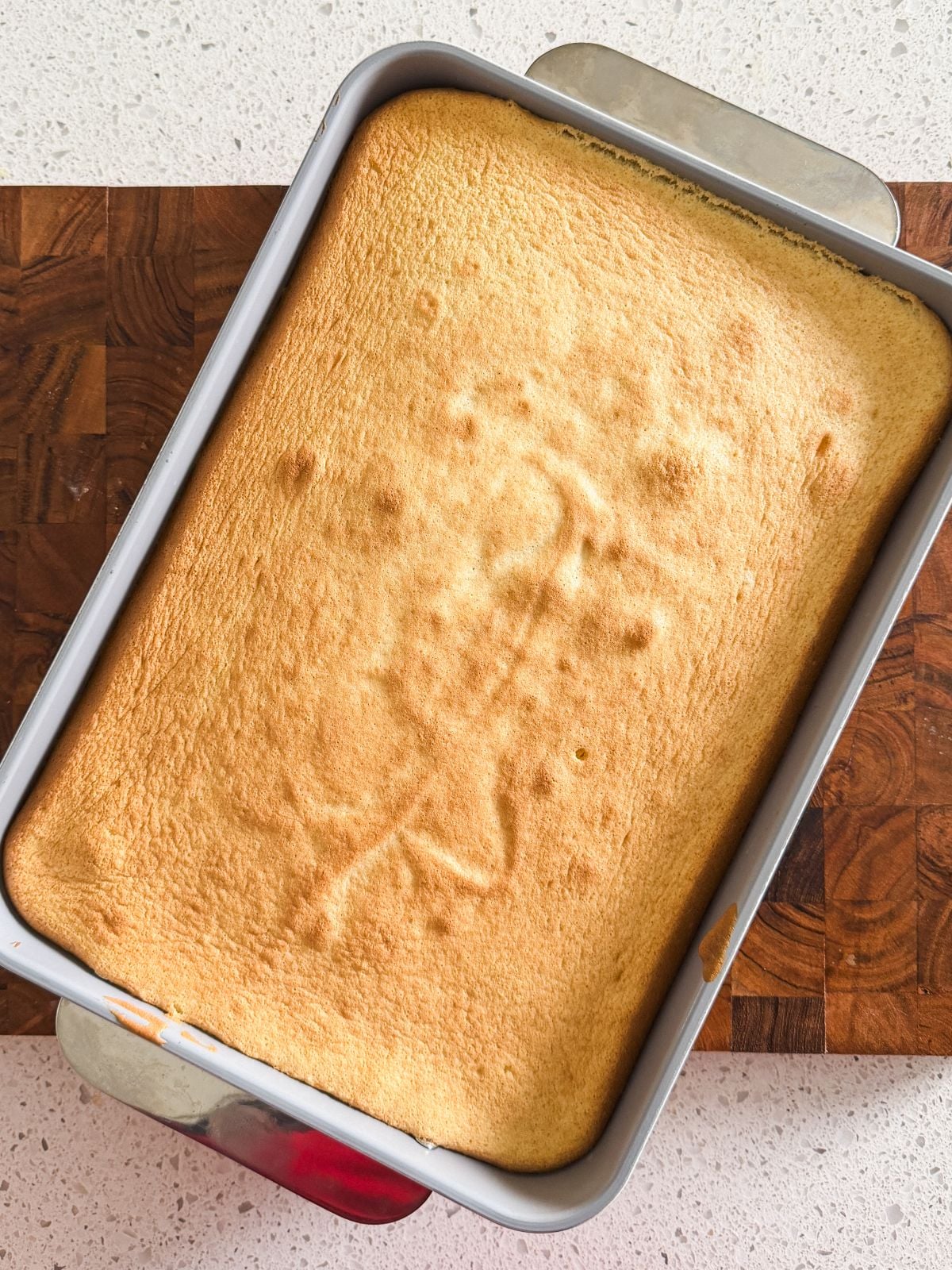 A rectangular golden-brown tres leches cake rests in a metal baking pan on a wooden cutting board and a white countertop. The cake's surface is smooth with a few small bumps.