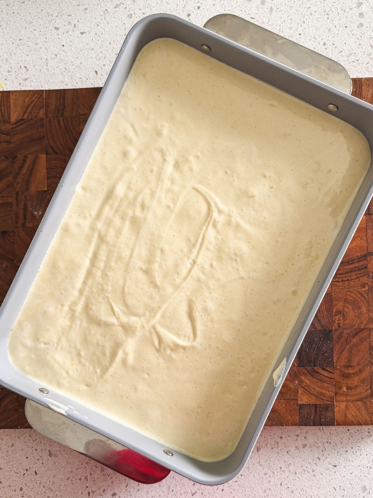 A rectangular metal baking pan filled with pale, smooth tres leches cake batter sits on a wooden cutting board atop a speckled countertop. Swirl marks are visible on the batter's surface.