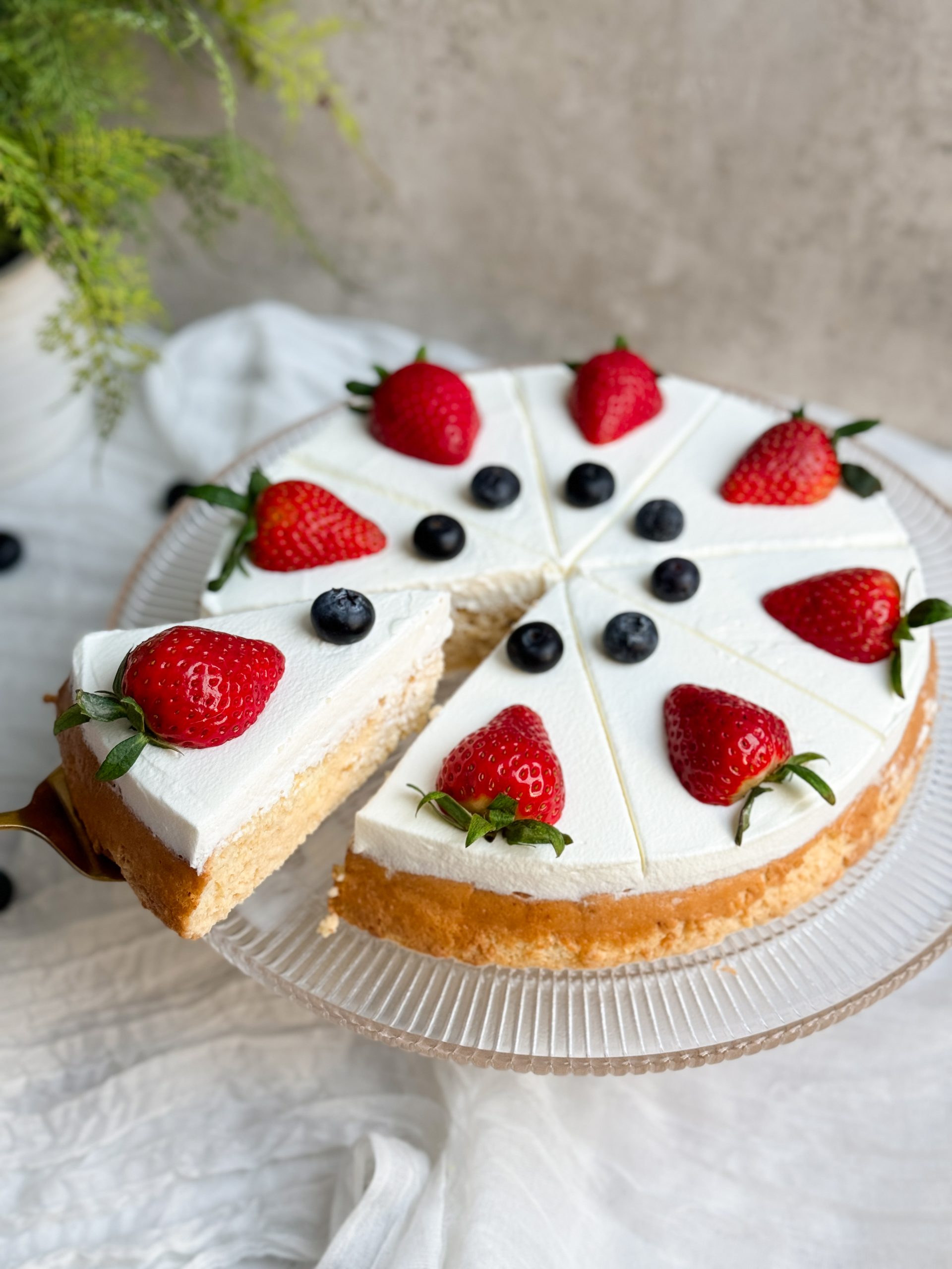 tres leches cake on a glass serving stand. cake is decorated with a thick layer of whipped cream and fresh berries. a spatula is pulling out a slice to show the moist and soft interior of the cake