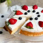 tres leches cake on a glass serving stand. cake is decorated with a thick layer of whipped cream and fresh berries. a spatula is pulling out a slice to show the moist and soft interior of the cake. close up pic