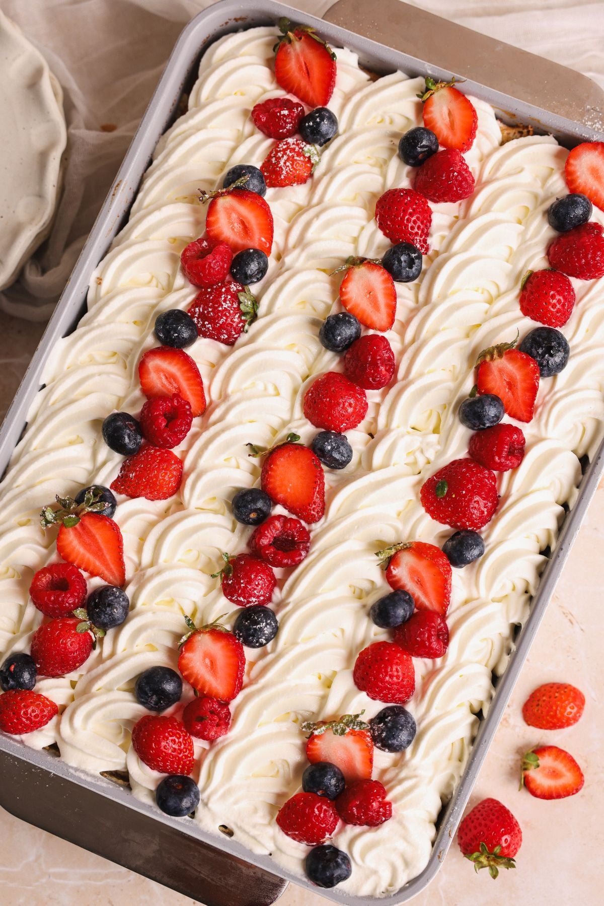 A rectangular tres leches cake topped with swirls of white whipped cream, decorated with rows of fresh strawberries, raspberries, and blueberries. The cake sits in a metal pan on a light surface with a few berries nearby.