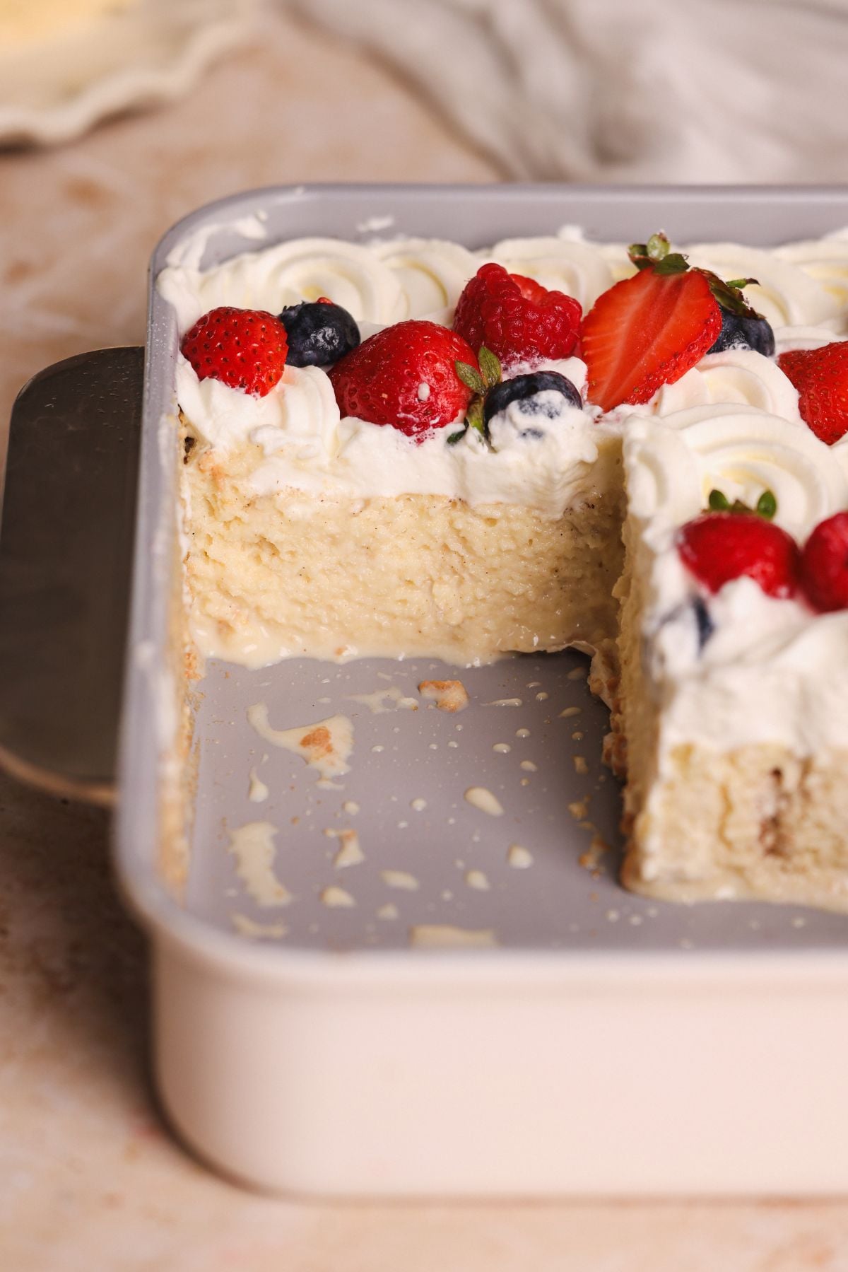 A rectangular tres leches cake with creamy frosting and mixed berries sits in a white baking dish. A large square piece has been cut out, revealing the cake’s moist layers. Whipped cream and berries decorate the top.