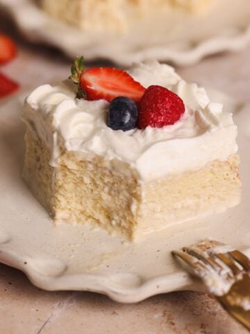 A decadent slice of tres leches cake topped with whipped cream, a strawberry half, a blueberry, and a raspberry sits on a scalloped plate with a gold fork, surrounded by scattered berries.