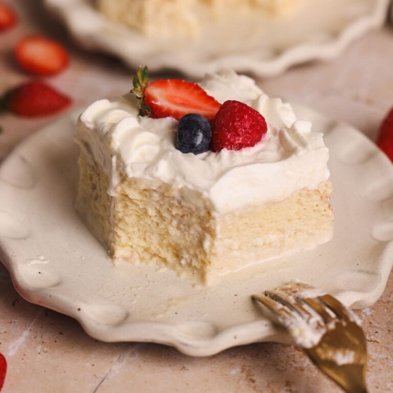 A decadent slice of tres leches cake topped with whipped cream, a strawberry half, a blueberry, and a raspberry sits on a scalloped plate with a gold fork, surrounded by scattered berries.