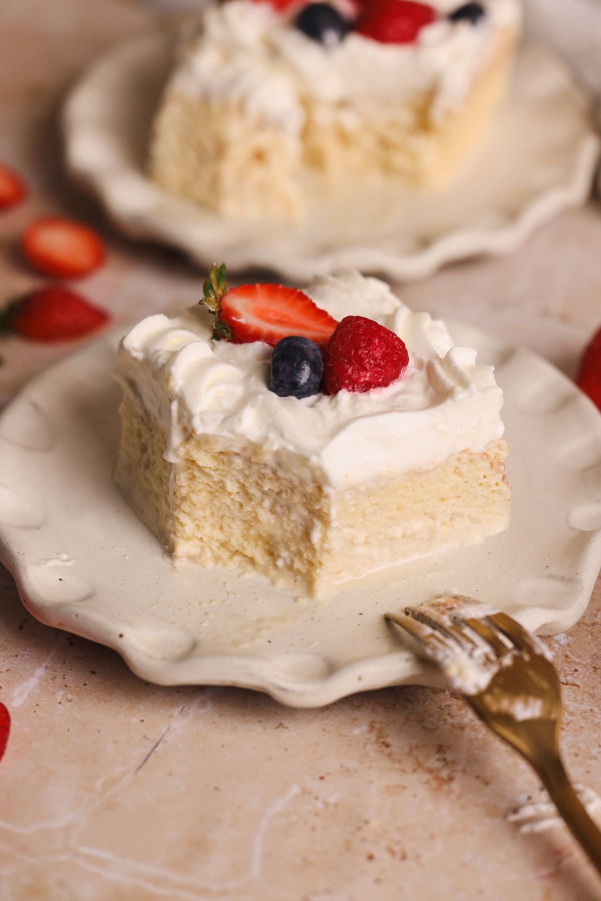 A square slice of creamy tres leches cake topped with whipped cream, a halved strawberry, a blueberry, and a raspberry sits on a decorative plate. A fork with cake remnants is nearby, and more fruit is scattered around.