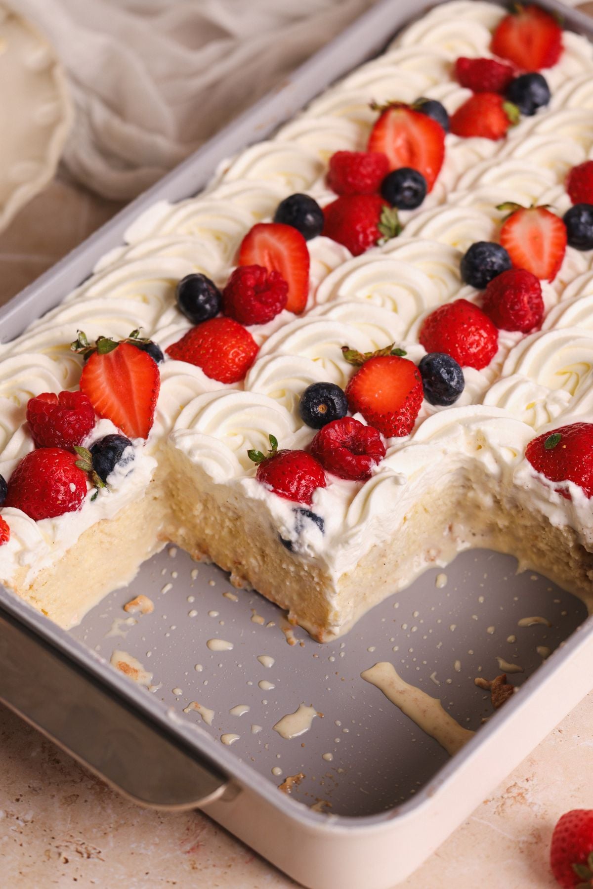 A rectangular tres leches cake topped with swirls of whipped cream and rows of fresh strawberries and blueberries sits in a baking pan with a few pieces missing.
