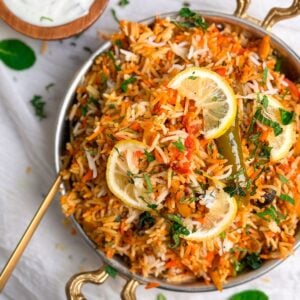 chicken biryani in a copper serving dish, garnished with lemon slices and mint leaves. A small bowl with yogurt raita on the side. partial picture from the top