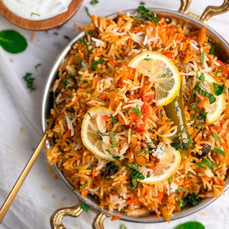 chicken biryani in a copper serving dish, garnished with lemon slices and mint leaves. A small bowl with yogurt raita on the side. partial picture from the top