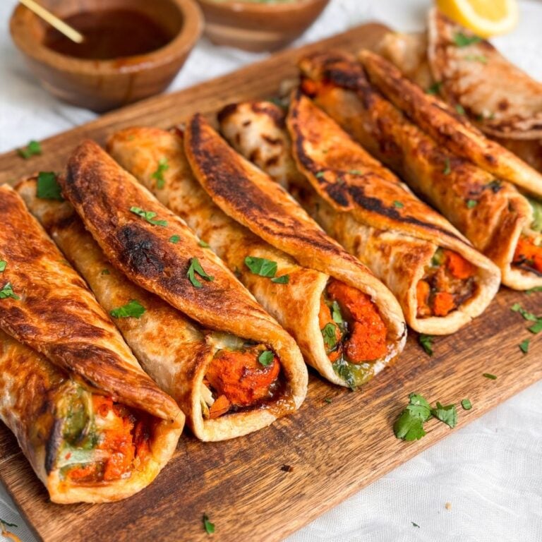 6 chicken tikka paratha rolls on a wooden cutting board garnished with cilantro. Rolls are golden and flakey and filled with red and juicy chicken tikka boti and chutneys. Picture from an angle to show the inside of the rolls