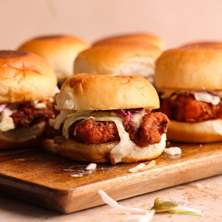 nashville hot chicken sliders on a wooden cutting board. one slider is in focus from the side showing crispy fried chicken, coleslaw and pickles inside the slider