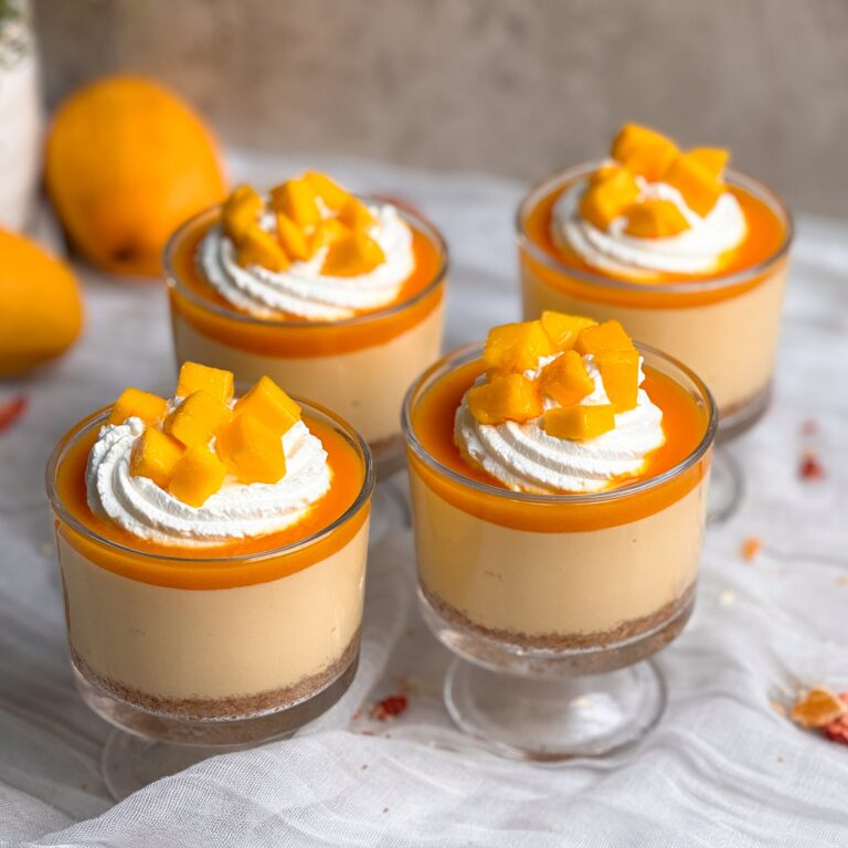 4 no bake mango cheesecake cups. Each cup has a layer of crust, mango cheesecake filling and mango pulp, and is deocrated with whipped cream and chopped mangoes. Mangoes seen in background