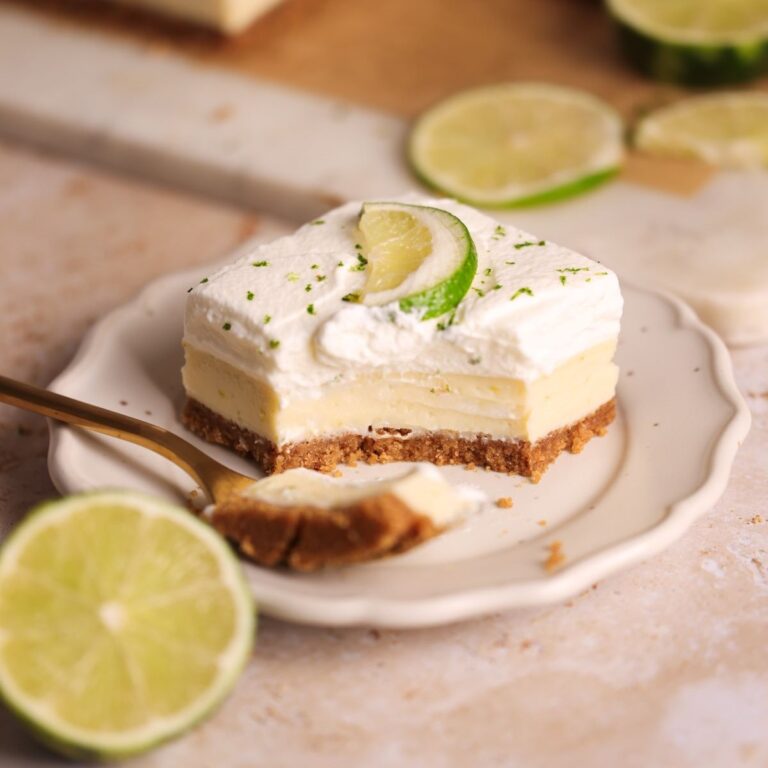 a key lime pie bar on a small plate with a fork taking out a bite and resting on the side. the bar has a graham cracker crust, a silky key lime interior, and whipped cream on top; decorated with lime zest