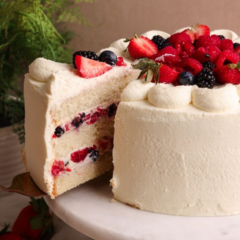 berry chantilly cake on a marble cake stand decorated with cream and berries, spatula pulling out a slice showing the soft texture of the cake and the cream and berry filling inside
