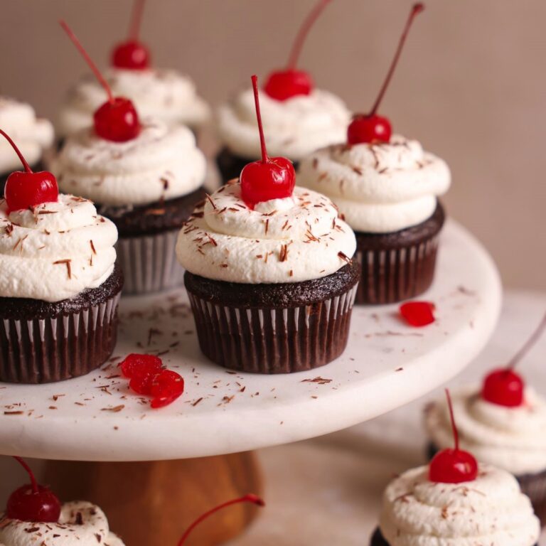 Black forest cupcakes on a white marble cake stand, with one in focus. The chocolate cupcakes are topped with whipped cream swirls and cherries