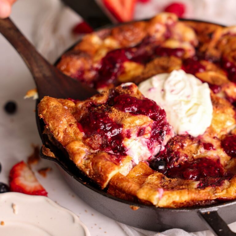 baked berry cheesecake french toast in a cast iron skillet with dollops of cheesecake and berry compote on top. a spatula is digging in, close up picture from the side