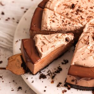 no bake chocolate cheesecake on a marble serving board. it is decorated with chocolate ganache dripping down the sides and chocolate whipped cream on top. spatula is pulling out a slice