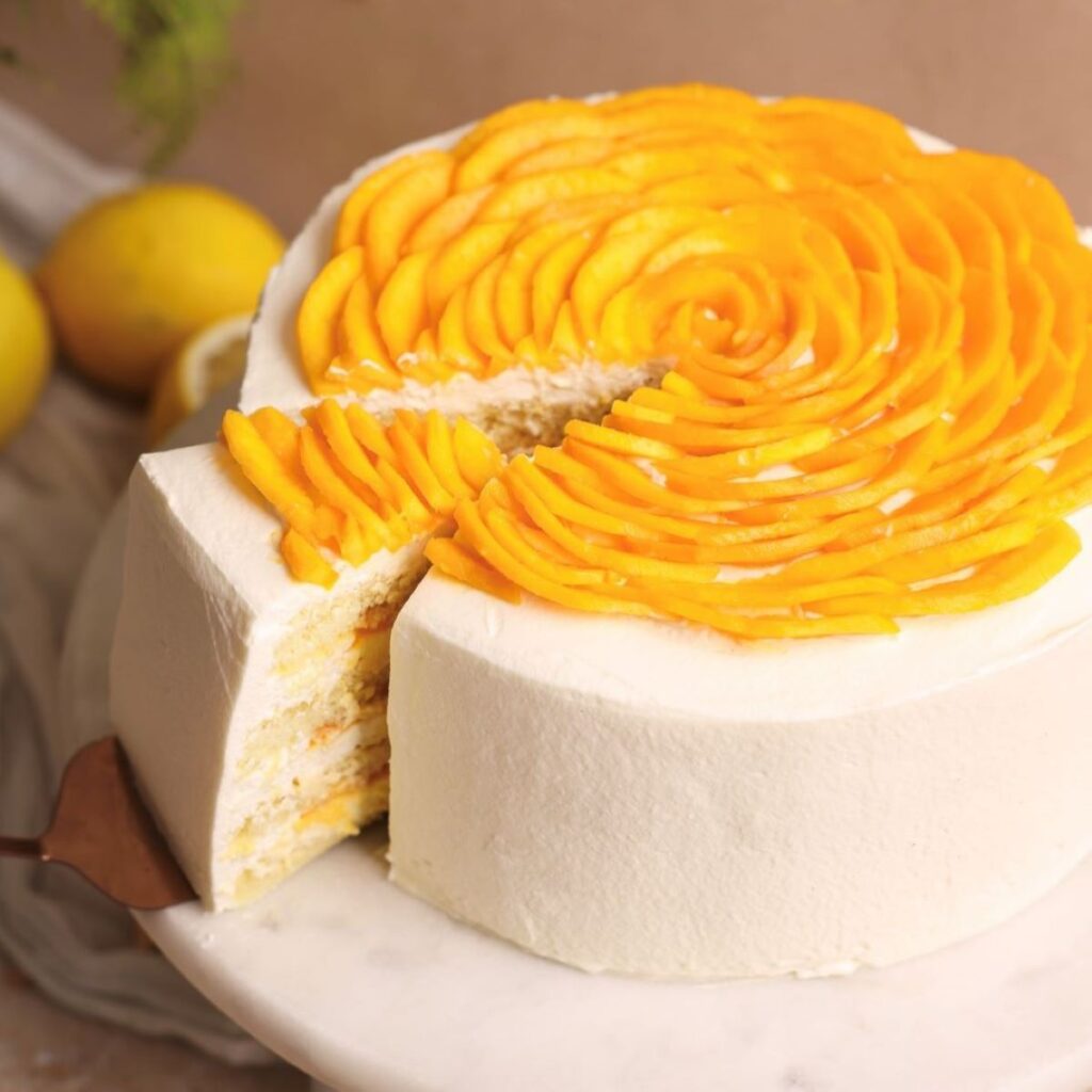 mango cream chiffon cake on a marble cake stand. cake is frosted with whipped cream and has sliced mangoes arranged like petals on top. A spatula is pulling out a slice showing the layers of cake, cream and mango inside