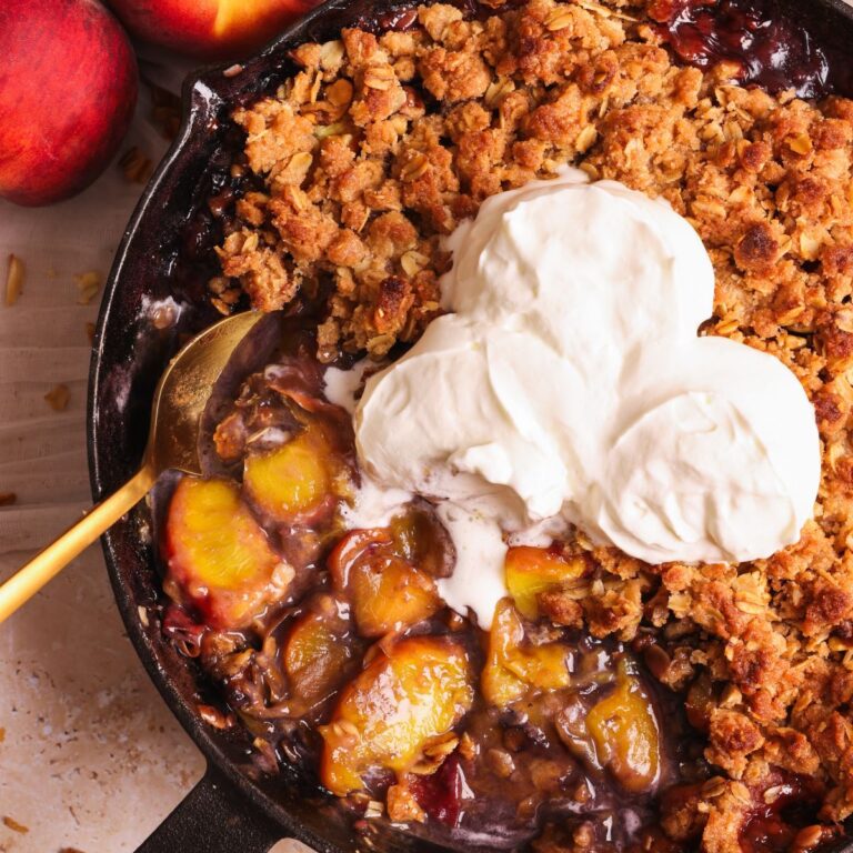 peach crumble in a cast iron skillet. The crumble is golden and crispy, topped with 3 scoops of whipped cream, and some of the crumble has been taken out to show the soft juicy peaches inside