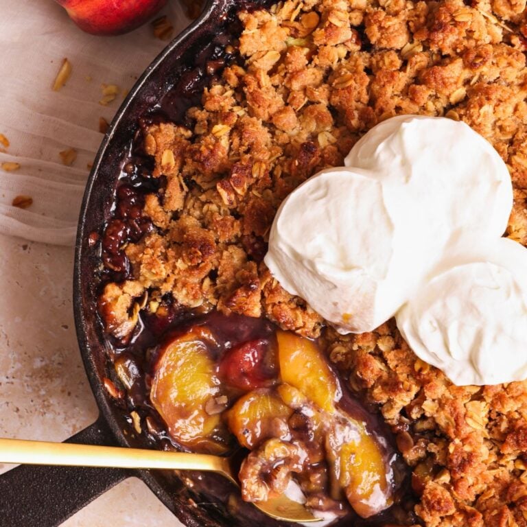 close up picture of peach crumble with a spoon in a cast iron skillet. The crumble is golden and crispy, topped with 3 scoops of whipped cream, and a serving has been taken out to show the soft juicy peaches inside