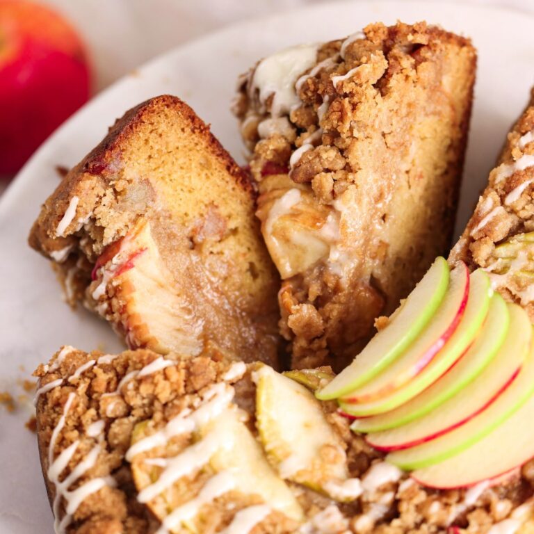 2 slices of apple crumb cake placed sideways next to the cake on a marble cake stand to show the soft moist cake, apples, and crumb in the cross-section