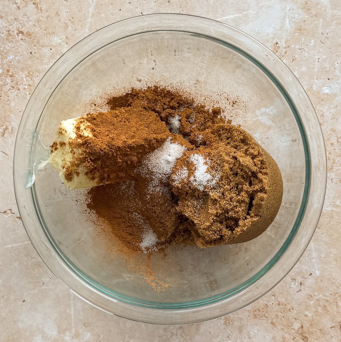 A glass bowl with butter, brown sugar, cinnamon, and a pinch of salt on a light countertop, seen from above