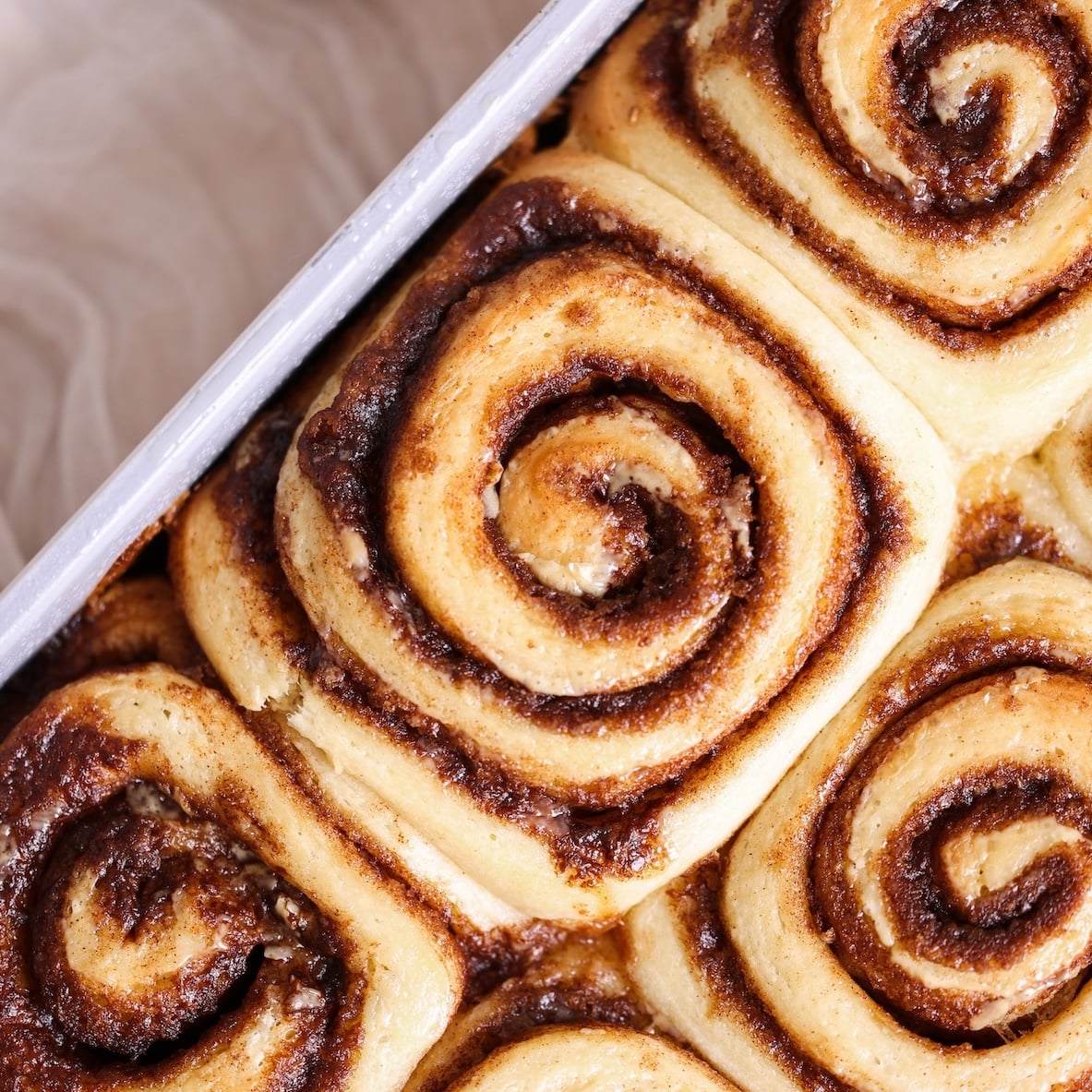 Close-up of freshly baked cinnamon rolls in a baking pan, with golden-brown swirls and a soft, fluffy texture. These cinnamon rolls are tightly packed, showcasing a rich cinnamon filling and slightly crisp edges.