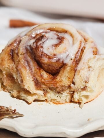 A close-up of one of a cinnamon roll with white icing drizzled on top, placed on a cream-colored plate. The roll looks soft and partially eaten, with a fork nearby.