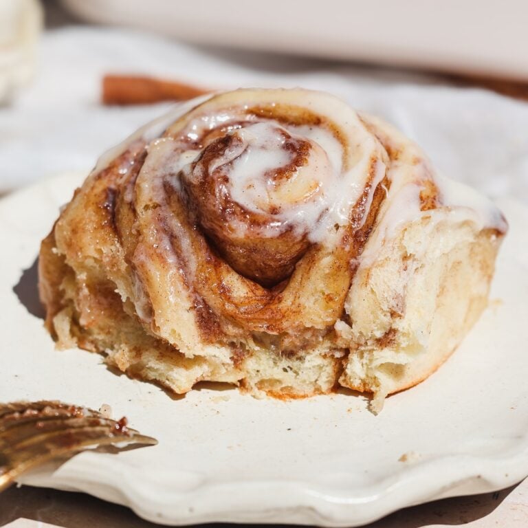 A close-up of one of a cinnamon roll with white icing drizzled on top, placed on a cream-colored plate. The roll looks soft and partially eaten, with a fork nearby.