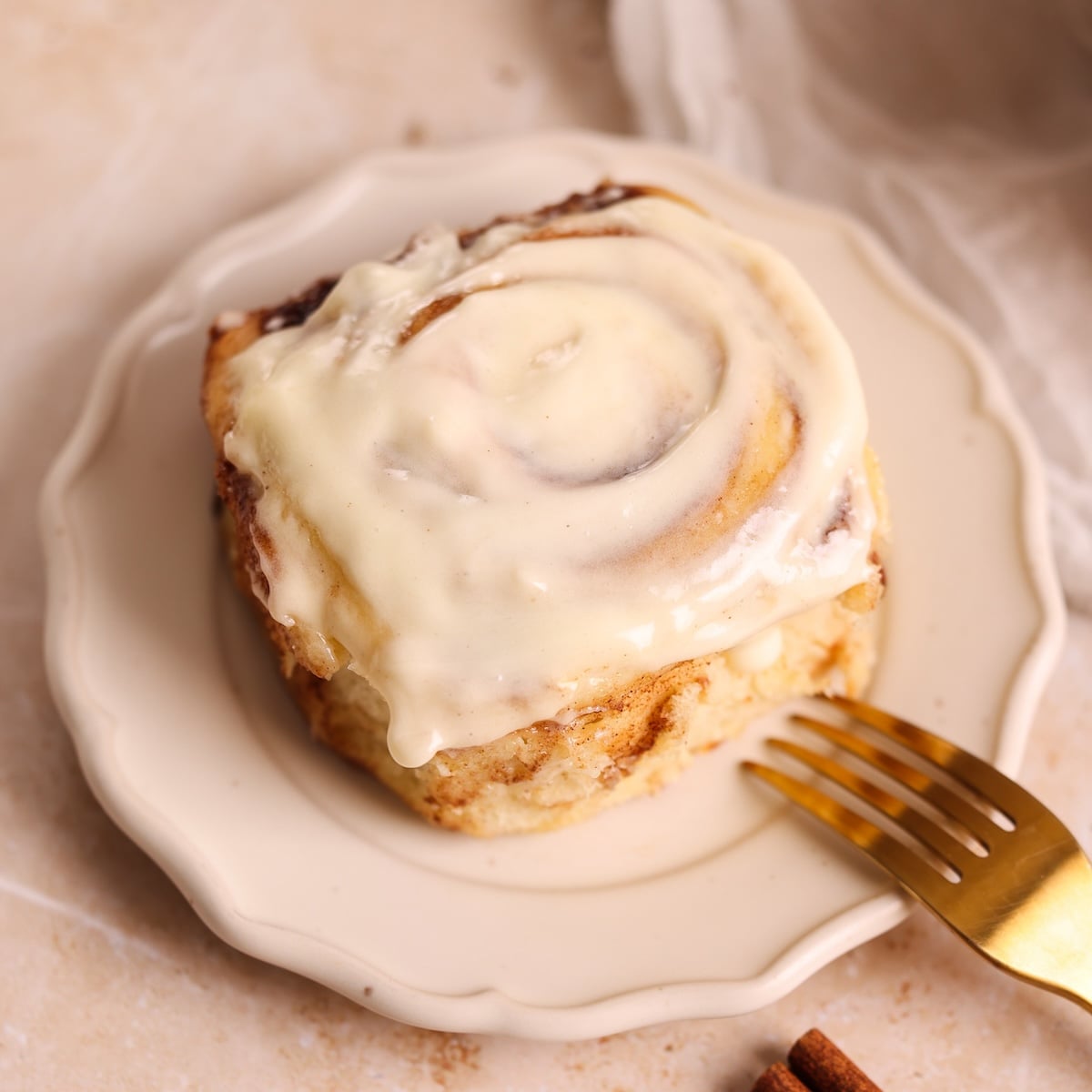 A cinnamon roll is topped with creamy white icing and sits on a small white plate with a gold fork beside it, against a light background.