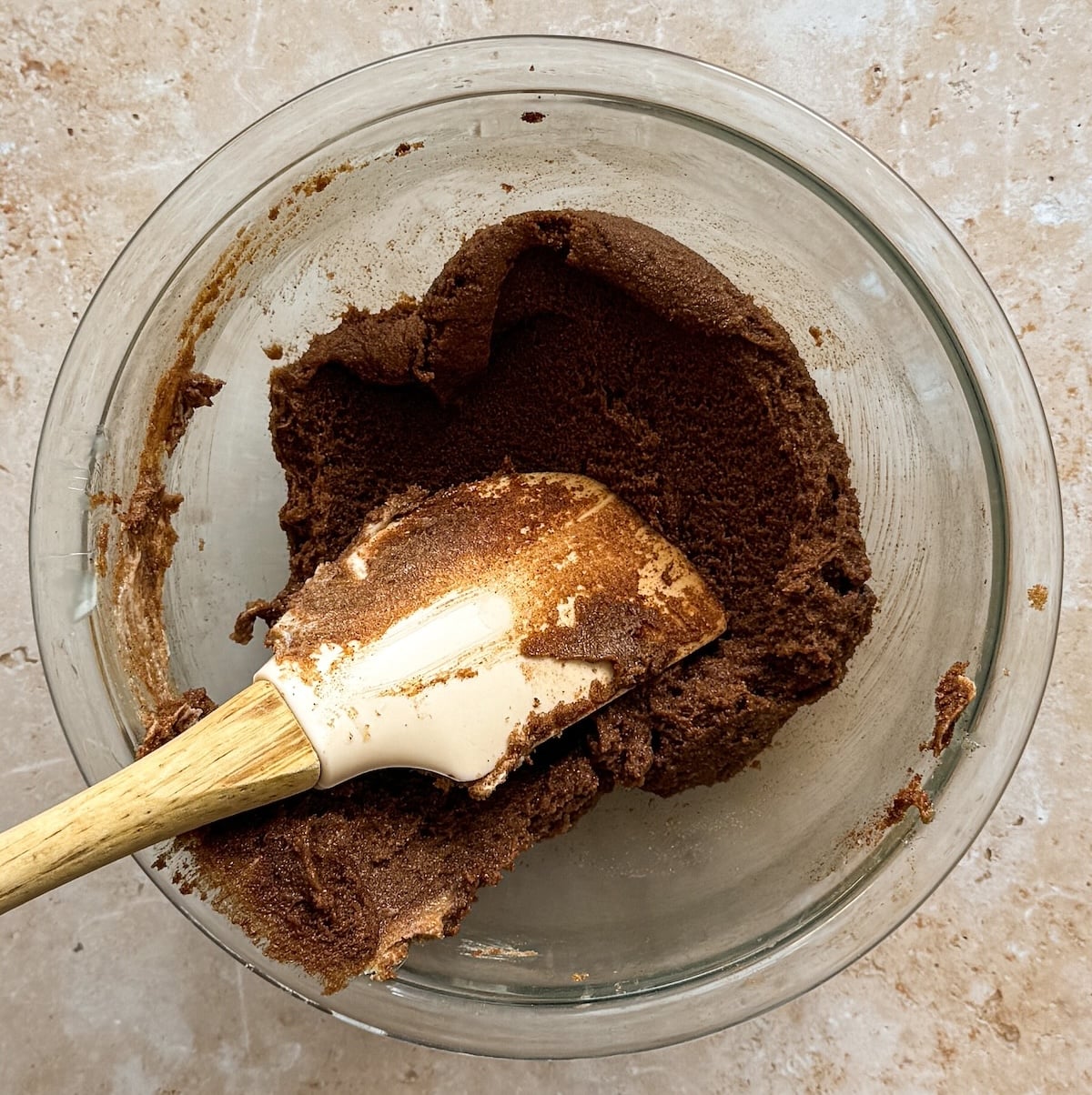 A glass bowl with a cinnamon sugar mixture being mixed by a white spatula with a wooden handle on a beige countertop