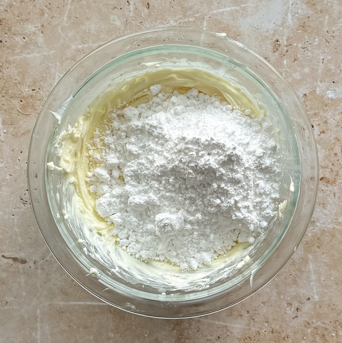 A glass bowl filled with partially mixed butter and a mound of powdered sugar, sitting on a beige countertop