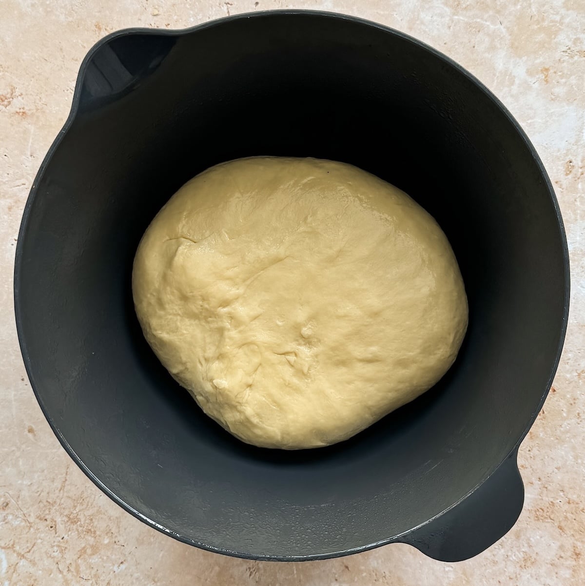 A ball of bread dough rests in a black mixing bowl on a light-colored surface, ready to rise 