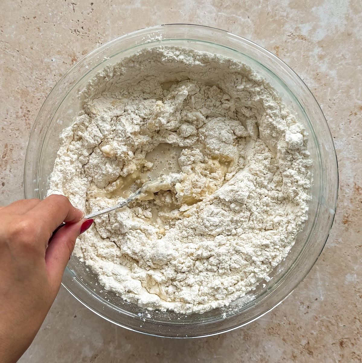 A hand stirs flour and liquid in a glass bowl with a metal spoon, beginning to mix dough on a beige countertop