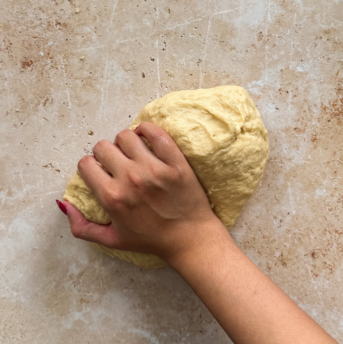 A hand kneads a ball of dough on a light-colored, lightly floured surface. the dough looks smoother than before