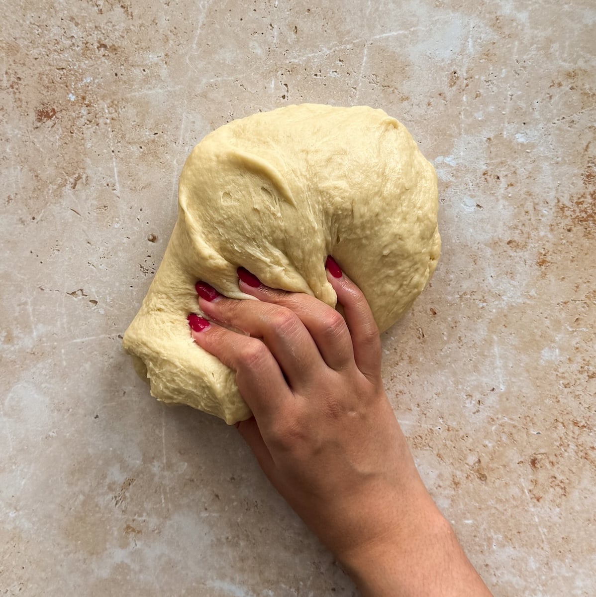 A hand with red nail polish kneads a soft, yellowish dough on a lightly floured, textured surface