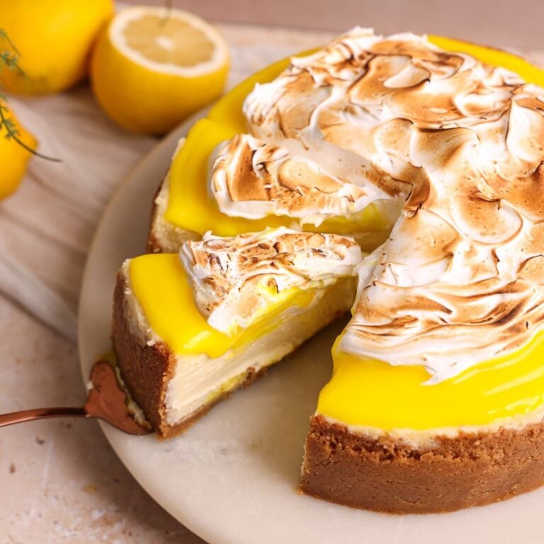 lemon meringue cheesecake on a marble cake stand. Cheesecake has a golden crust and is topped with lemon curd and torched meringue, with a spatula pulling out a slice
