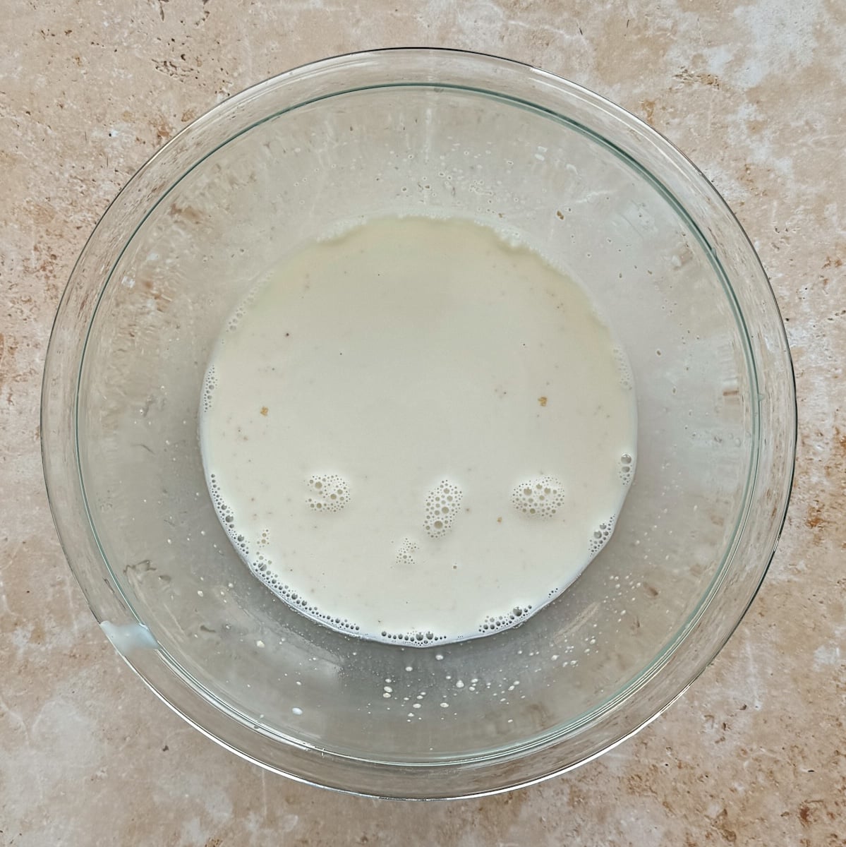 A glass bowl with a small amount of foamy, off-white liquid sits on a tan, speckled countertop