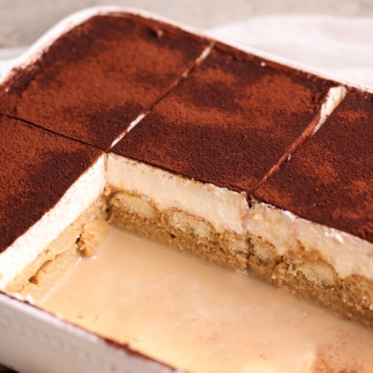 tiramisu tres leches in a rectangular dish dusted with cocoa powder and cut into 8 slices. 2 slices have been removed to show the layers of soaked ladyfingers and mascarpone whipped cream. close up picture from an angle