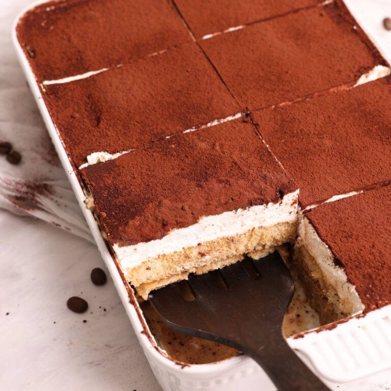 spatula pulling out a slice of tiramisu tres leches from the dish; showing layers of soaked sponge, mascarpone whipped cream and cocoa powder on top