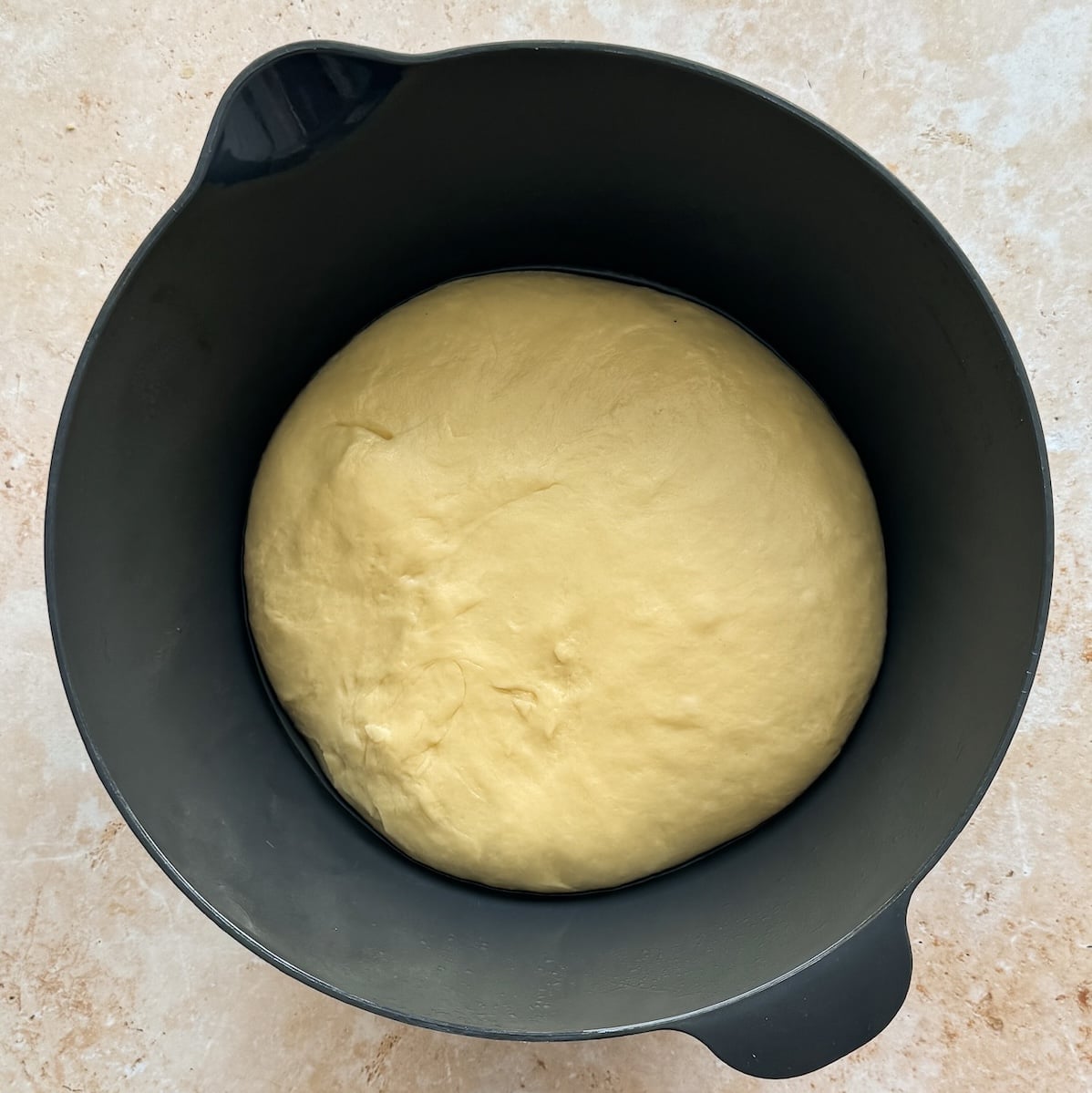 A smooth, risen ball of dough sits in a black mixing bowl on a light-colored countertop.
