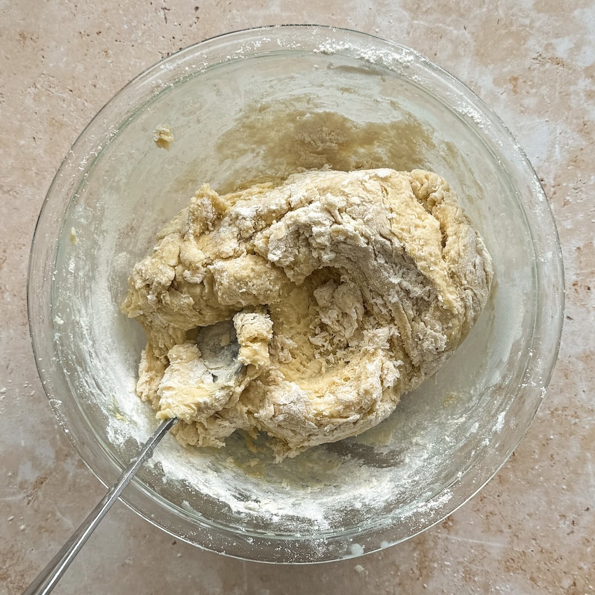 A glass bowl with partially mixed dough and a spoon on a light, flour-dusted surface.