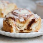 A close-up of a cinnamon roll topped with creamy white icing sits on a white plate, with another cinnamon roll blurred in the background. It looks soft and gooey inside