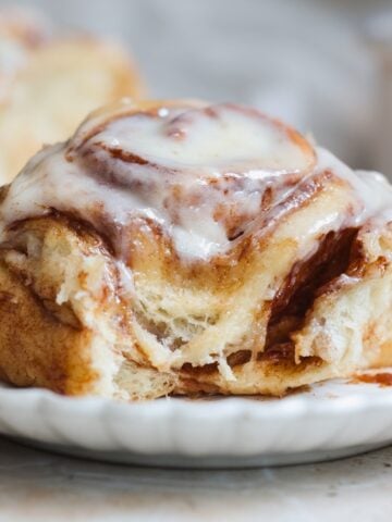 A close-up of a cinnamon roll topped with creamy white icing sits on a white plate, with another cinnamon roll blurred in the background. It looks soft and gooey inside